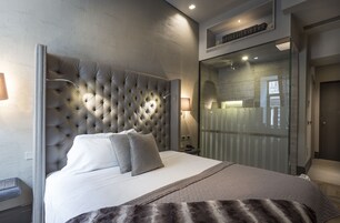 Un dormitorio moderno con cabecera capitonada, una cama grande, un box de ducha de vidrio y una mesita de noche con lámpara.