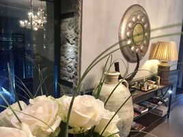 Una habitación con pared de piedra, un espejo, un jarrón con flores, una lámpara y una estantería.