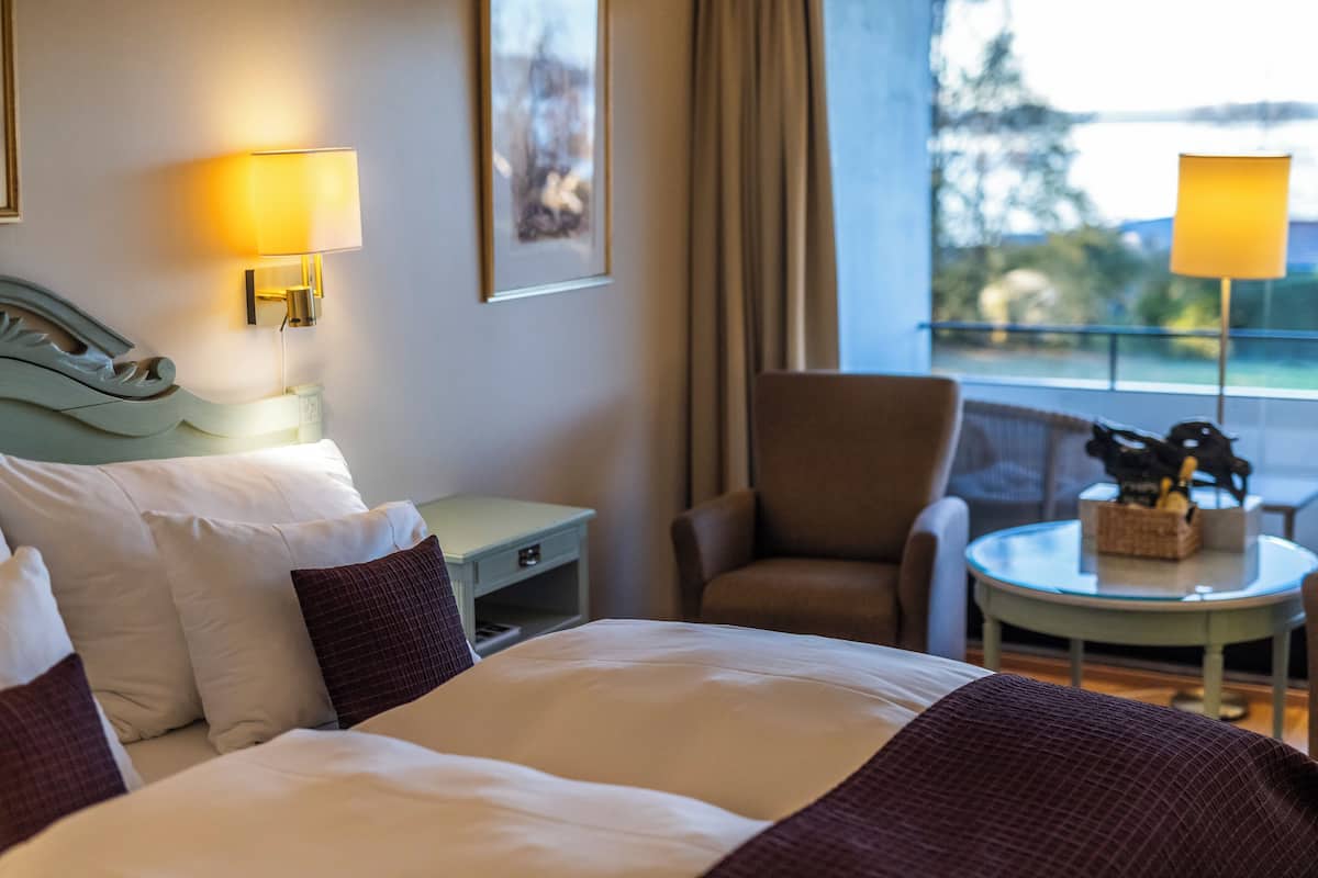 Une chambre d’hôtel avec un lit, deux lampes de chevet, une chaise, une petite table avec une bouteille et une fenêtre avec des rideaux.