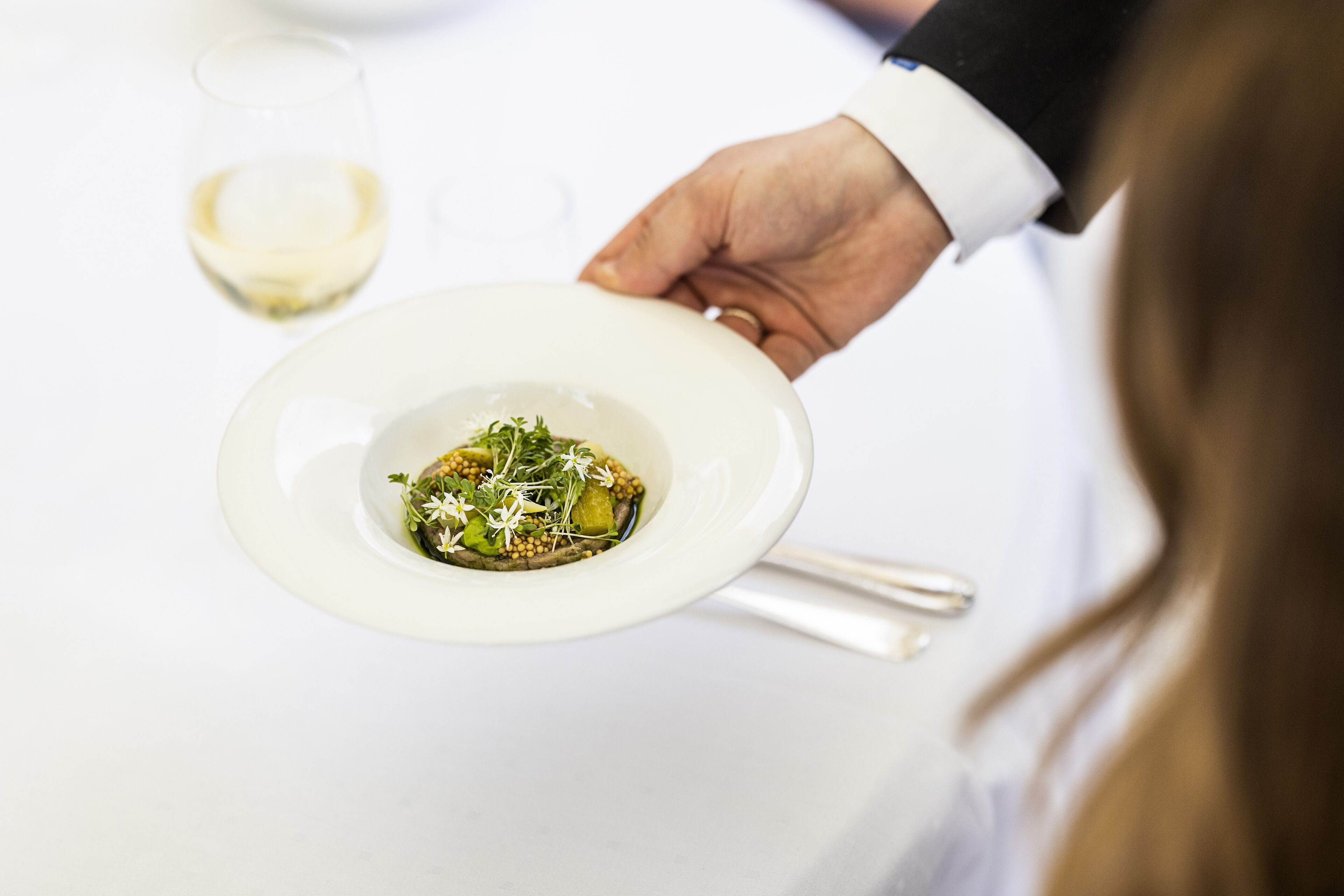 A person holding a white plate with a dish of food, a glass of white wine, and a fork on a white table.