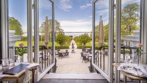 Une vue depuis une pièce dotée de grandes fenêtres donnant sur un jardin avec des tables et des chaises, une statue et un ciel dégagé.