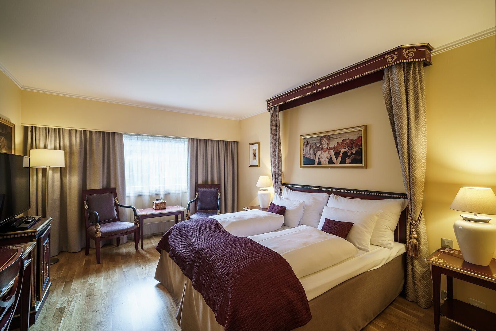 Habitación de hotel con cama con dosel, un escritorio con silla, televisor y un cuadro en la pared.