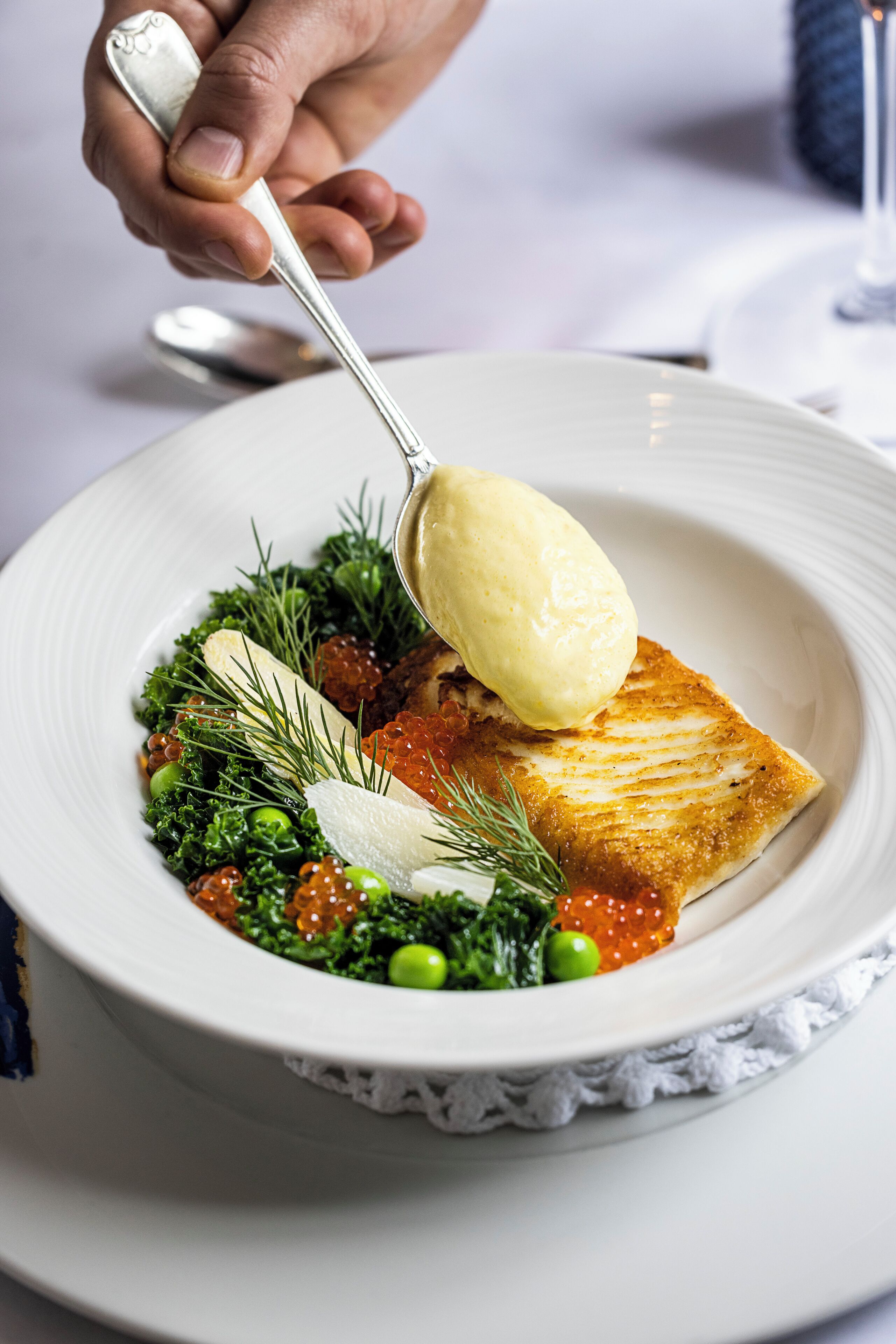 A white plate with a piece of fish, greens, and roe, and a spoon with a scoop of sauce.