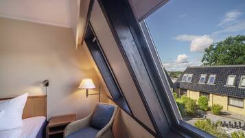 Una habitación con cama, una silla, una mesita de noche, una lámpara y una ventana con vista a casas y árboles.