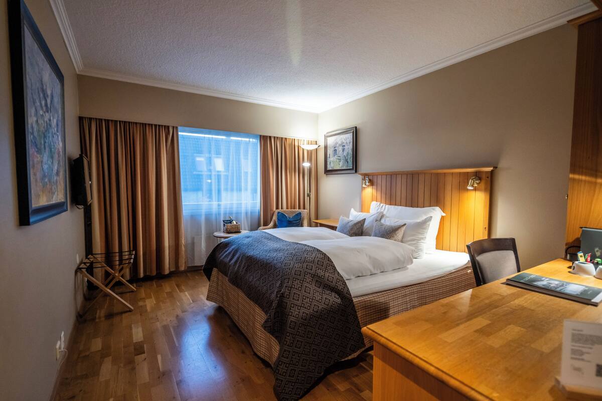Une chambre d’hôtel avec un grand lit, un bureau, une chaise, une lampe et une fenêtre avec des rideaux.