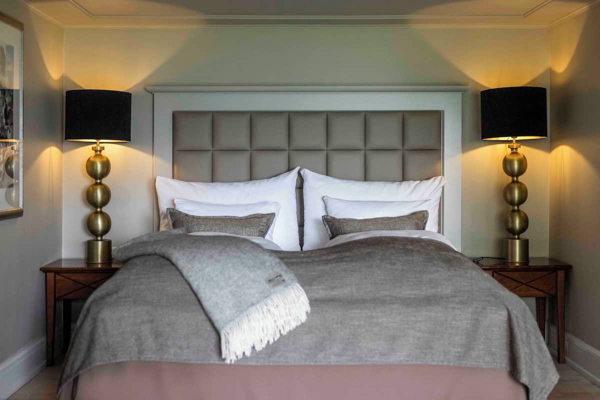Une chambre à coucher avec une tête de lit capitonnée, deux tables de chevet avec des lampes et un lit recouvert d’une couverture et de coussins gris.