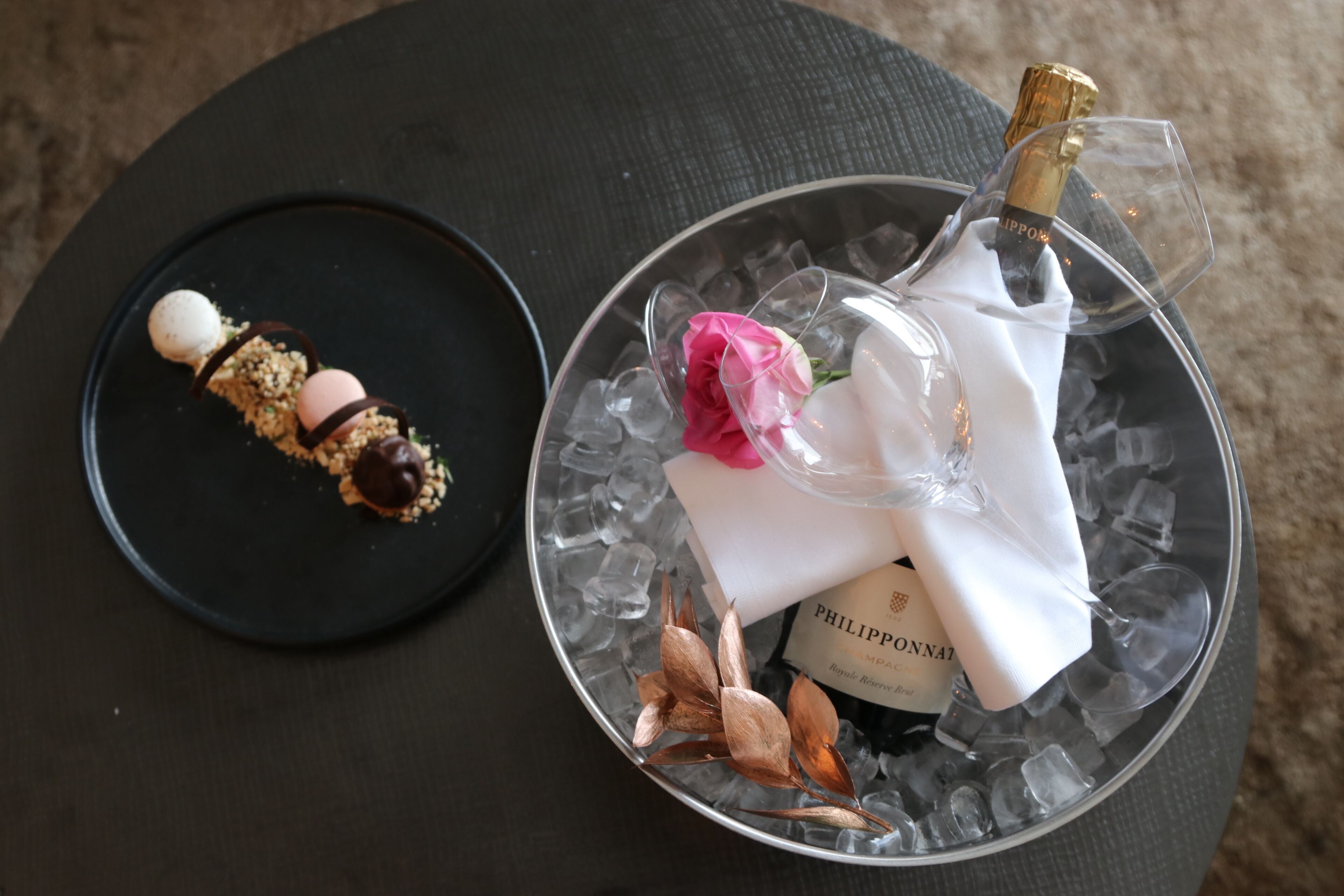 Un postre sobre un plato negro y una botella de champán con cubitos de hielo y una rosa sobre una mesa redonda.