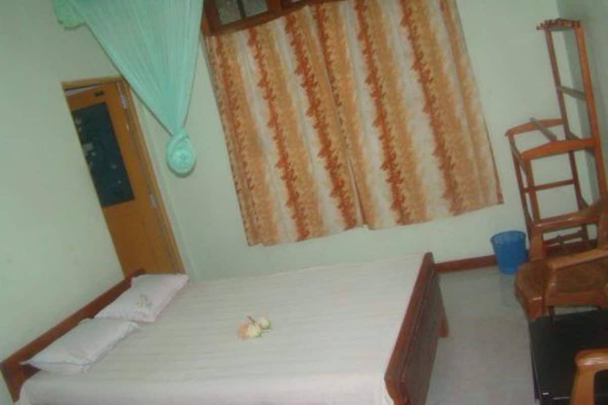 A single bed with a white bedspread and pillows, a wooden chair, a small table, a window with striped curtains, and a wooden door.