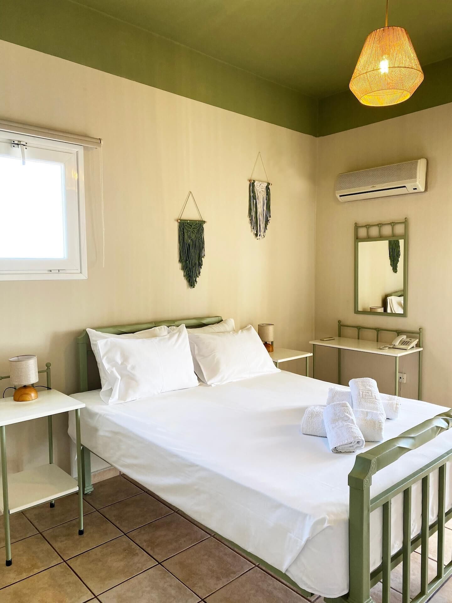 A hotel room with a bed, bedside tables, a mirror, an air conditioning unit, and wall-mounted macramé decorations.