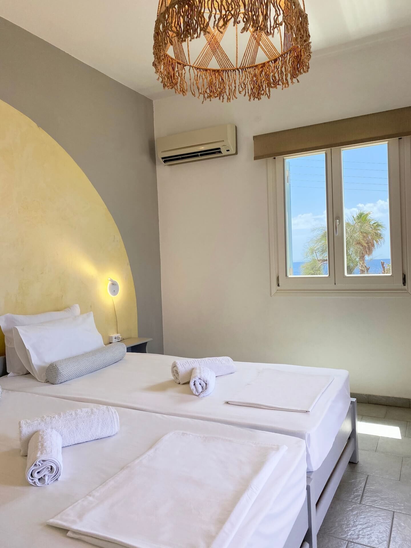 A hotel room with two beds, a large window with a view, an air conditioning unit, and a unique woven light fixture.