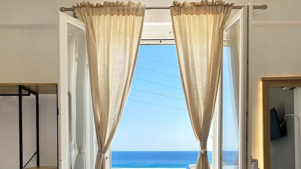 Quarto com cama, mesinha de cabeceira de madeira, mesa redonda pequena com cadeiras e vista para o mar através de cortinas abertas.