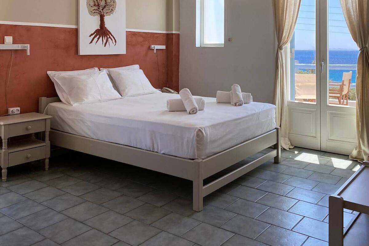 A bedroom with a bed, nightstands, a painting, a window, and a view of the sea.