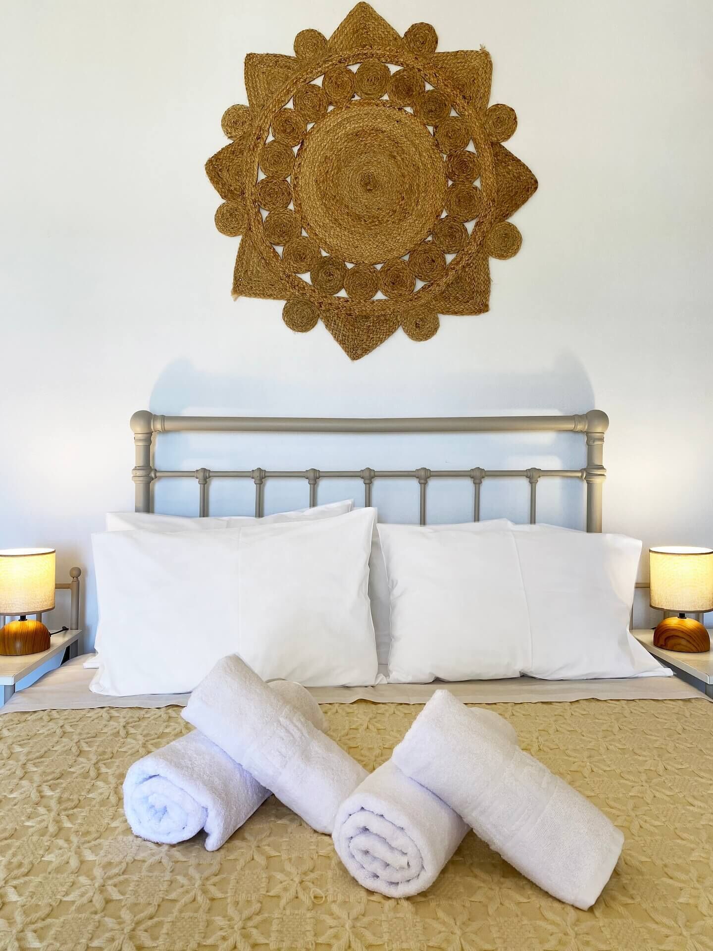 A neatly made bed with white linens and rolled towels, a decorative woven wall hanging above, and bedside lamps on either side.