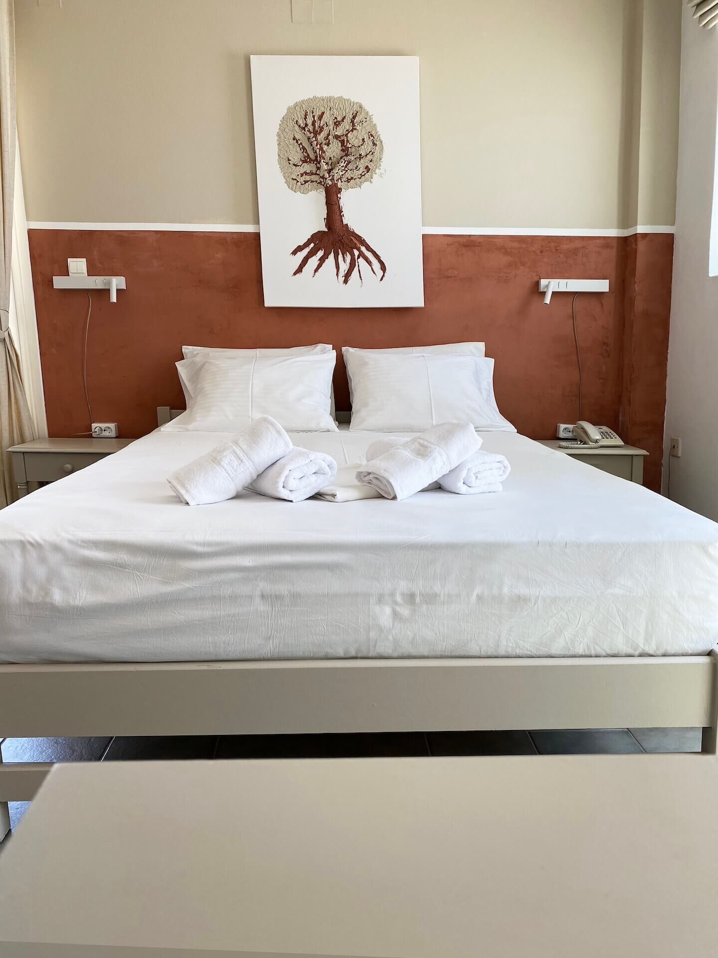Ein ordentlich bezogenes Bett mit weißen Bettwäsche und Handtüchern, ein an der Wand befestigtes Kunstwerk mit einem Baum und ein Nachttisch mit Telefon und Lampe.
