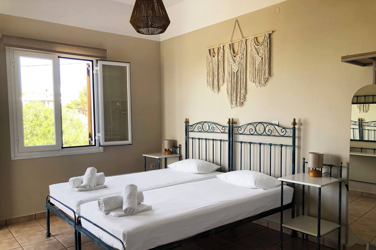A double bed with white linens and towels, a bedside table with a lamp, a small table with a lamp, a window with a view of greenery, and a macrame wall hanging.
