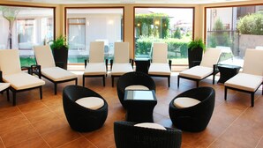 Amplia zona de estar interior con sillones y mesas, y ventanas grandes que ofrecen vistas al jardín exterior y edificios cercanos.
