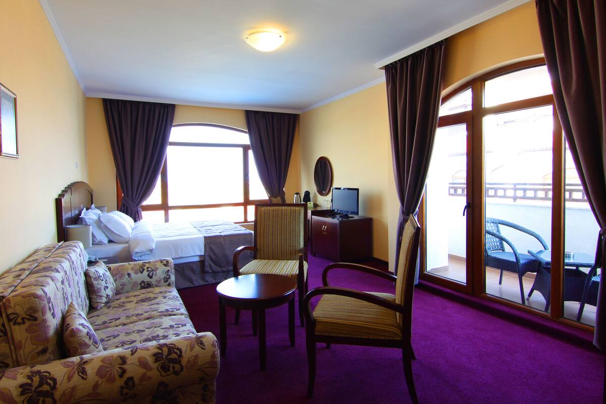A hotel room with a bed, a sofa, a chair, a desk with a computer, and a window with curtains.