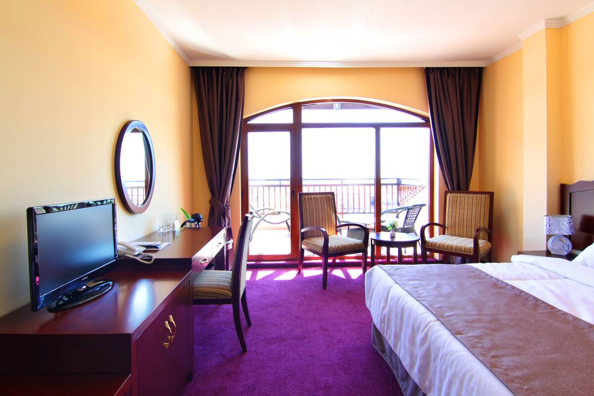 A hotel room with a bed, a desk with a TV, a chair, a table, and a balcony view.