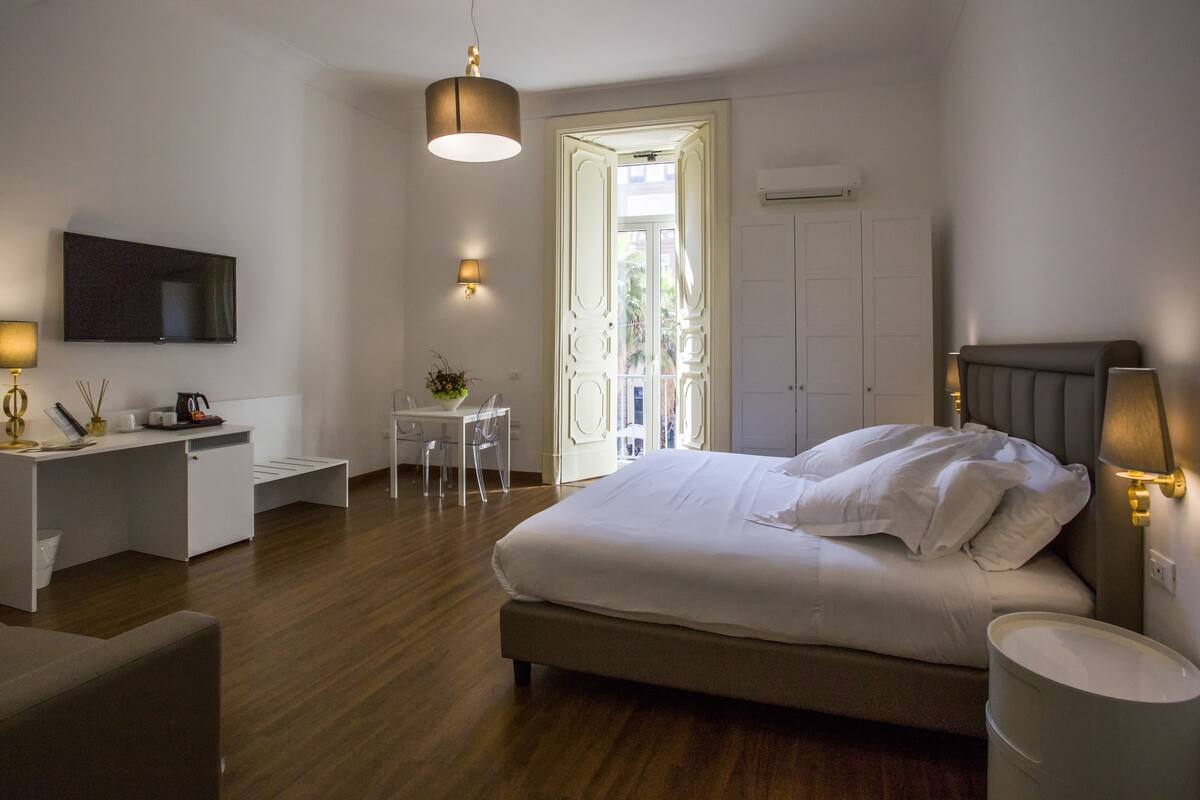 A modern hotel room with a large bed, a desk with a chair, a TV mounted on the wall, and a balcony with a view.