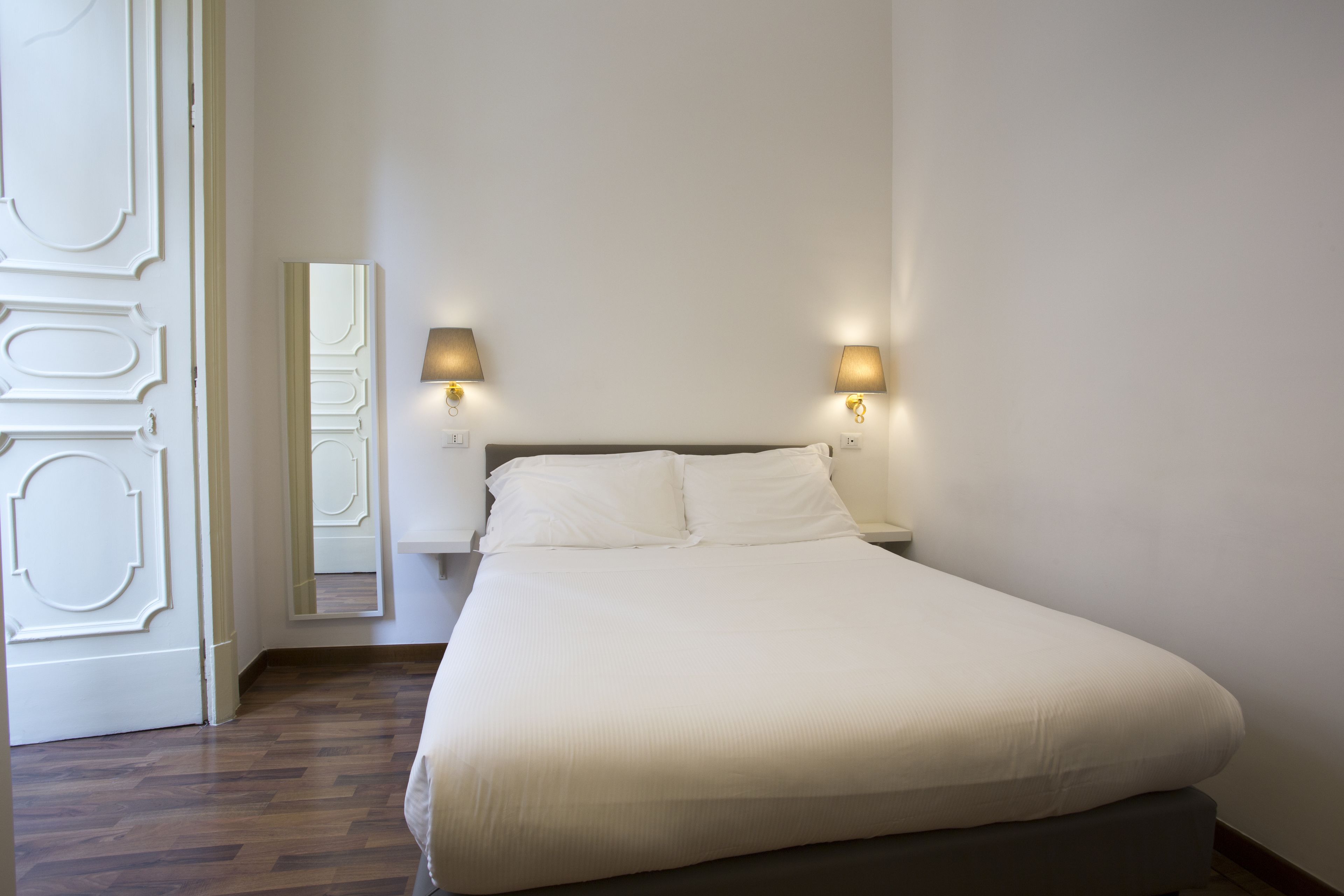 A neatly made bed in a room with a wooden floor, two bedside lamps, and a mirror on the wall.