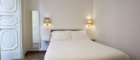 A neatly made bed in a room with a wooden floor, two bedside lamps, and a mirror on the wall.