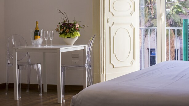 A hotel room with a white bed, a dining table with a bottle of champagne and glasses, a clear chair, and a potted plant. - Napoli Class Luxury Rooms & Suites (Naples)