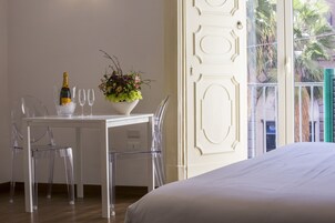 Una camera d'albergo con un letto bianco, un tavolo da pranzo con una bottiglia di champagne e dei bicchieri, una sedia trasparente e una pianta in vaso.