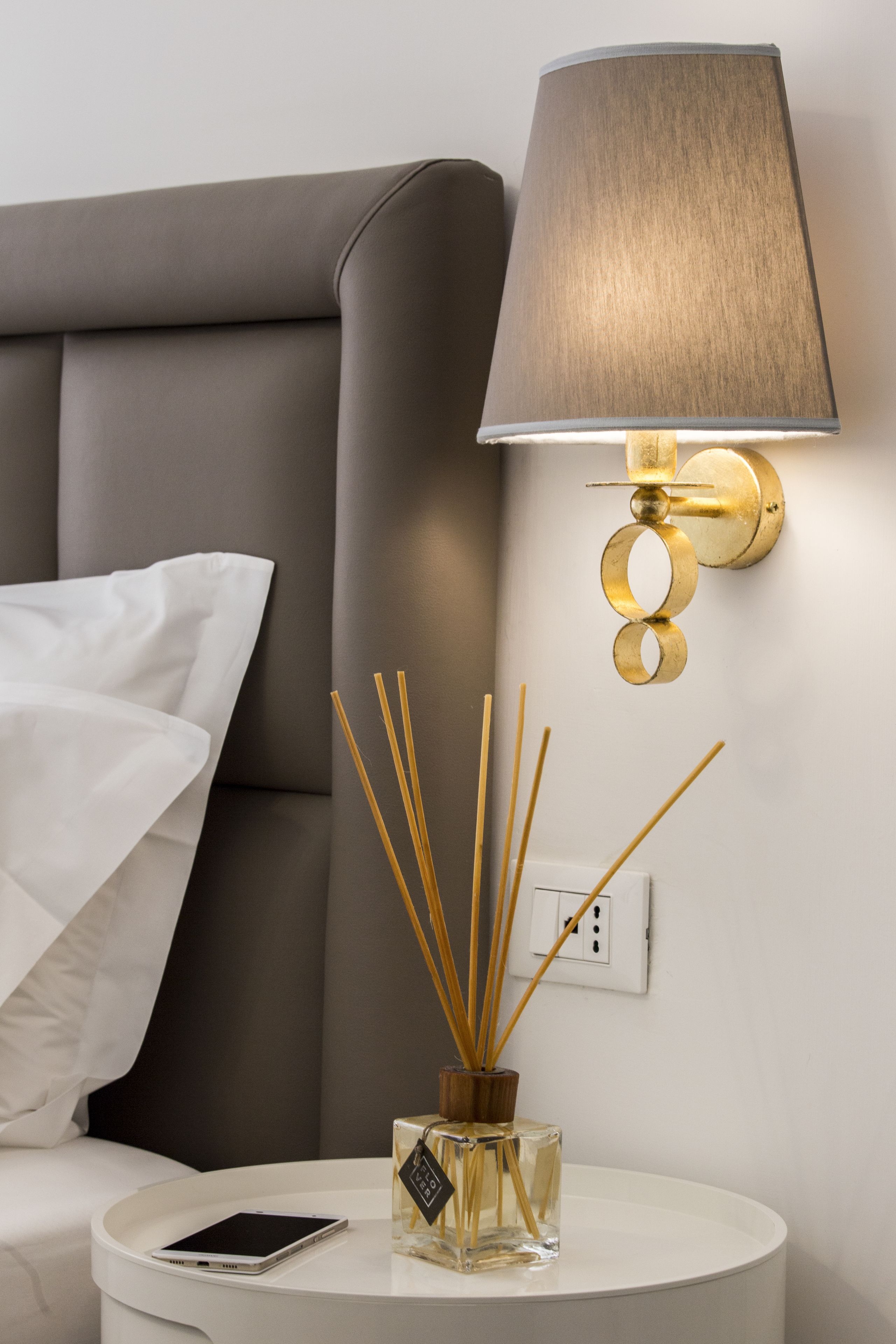 A modern room with a grey upholstered headboard, a round bedside table with a smartphone and a glass bottle of reed diffuser, and a wall-mounted lamp with a gold finish.