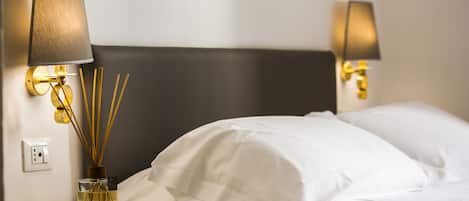 A neatly made bed with white linens, a bedside table with a lamp and a glass bottle of liquid, and a wall socket.