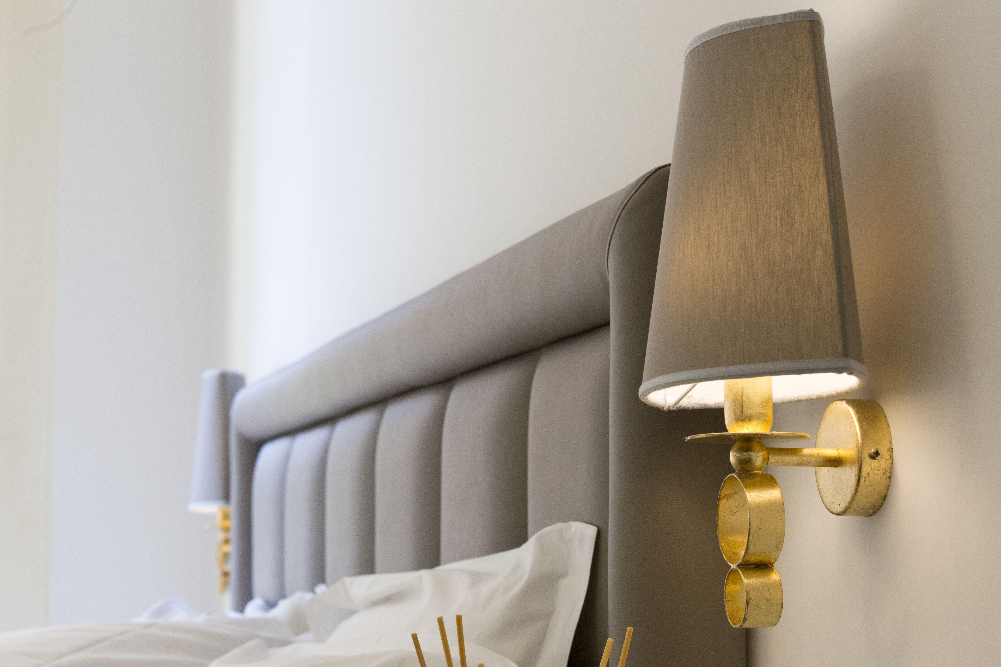 A bed with a grey upholstered headboard and a wall-mounted lamp with a cylindrical shade.