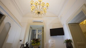 A reception area with a chandelier, a flat-screen TV, a potted plant, and a sign with text. - Napoli Class Luxury Rooms & Suites (Naples)