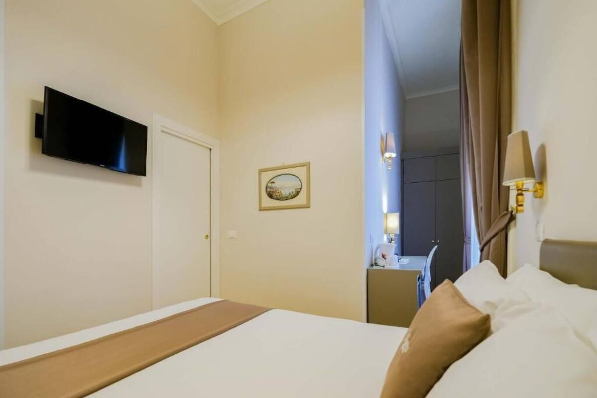 A hotel room with a bed, a television mounted on the wall, a small table with a lamp, and a picture frame on the wall.