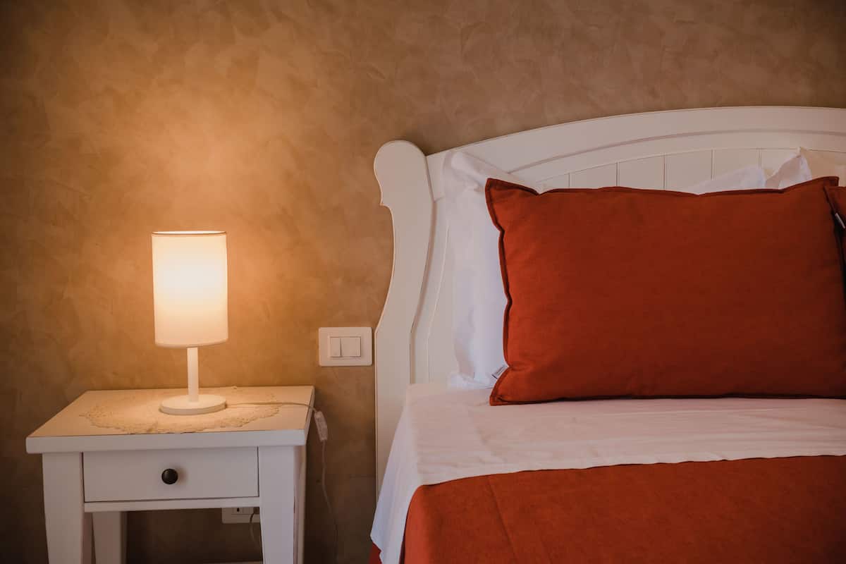 A neatly made bed with a white headboard and a red pillow, a bedside table with a lamp, and a textured wall.