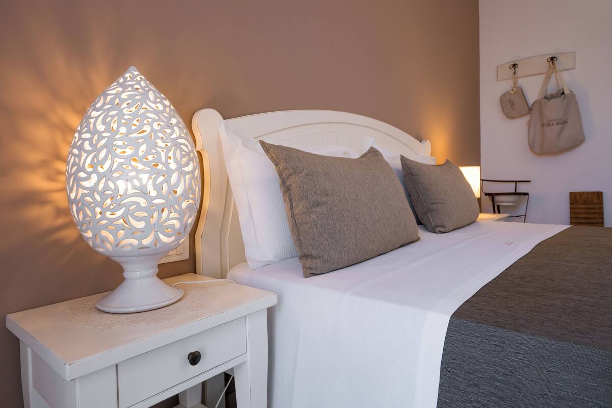A neatly made bed with a white headboard, a bedside table with a decorative lamp, and a wall-mounted bag rack.