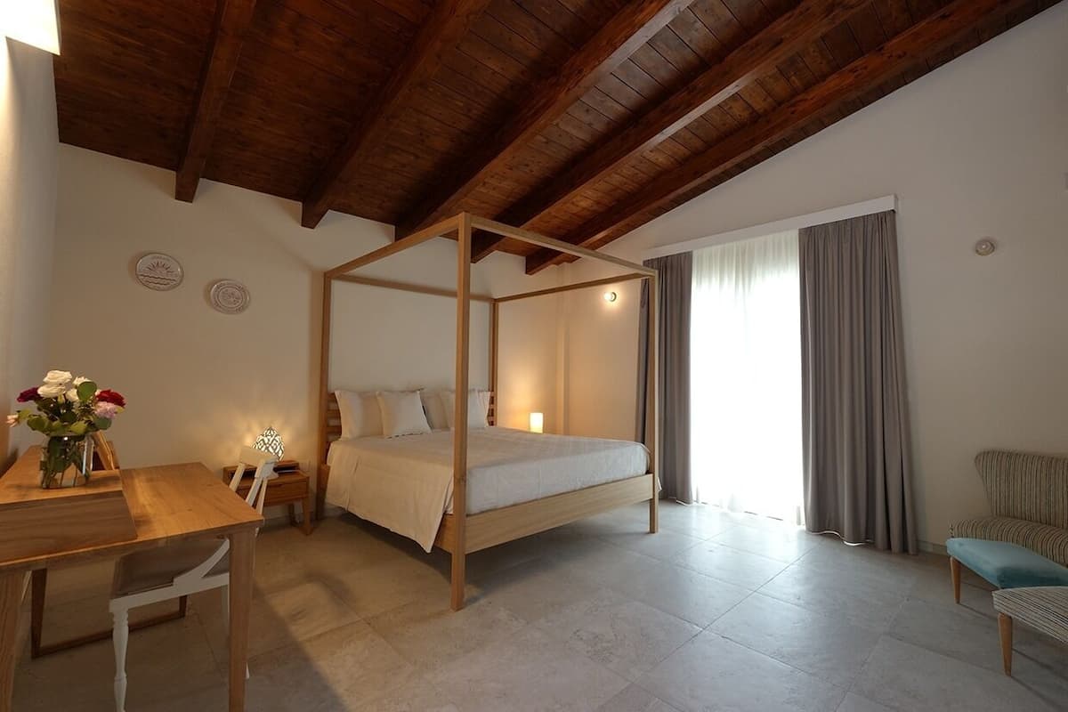 A bedroom with a wooden four-poster bed, a wooden desk with a chair, a small table with flowers, and a window with curtains.
