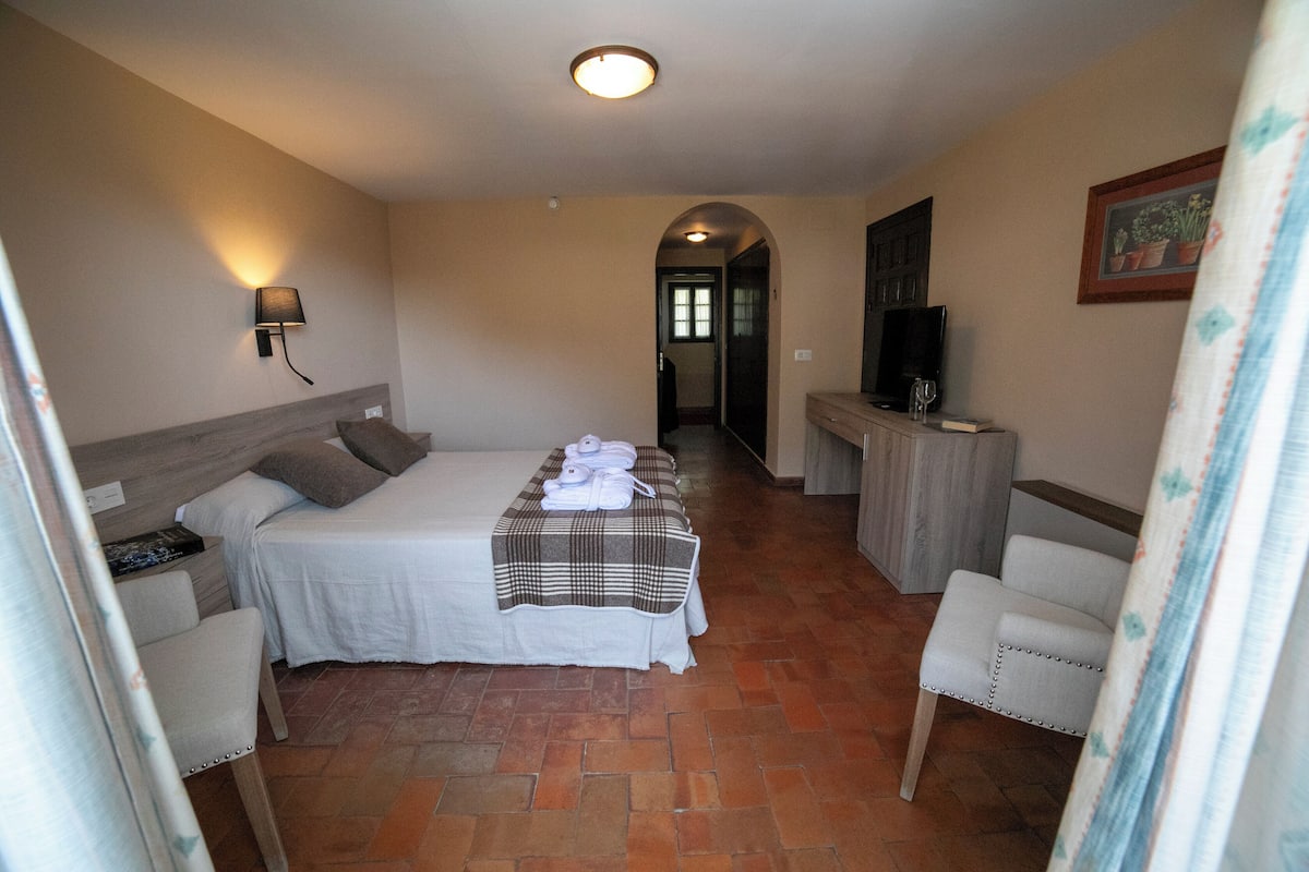Habitación de hotel con cama, escritorio, televisor y baño visible a través de un arco.