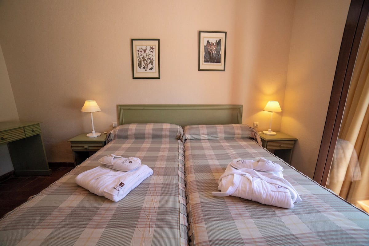 Habitación de hotel con dos camas, cada una con almohada y lámpara sobre una mesita de noche. Hay cuadros enmarcados en la pared.