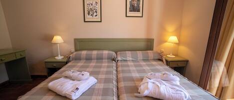 Habitación de hotel con dos camas, cada una con almohada y lámpara sobre una mesita de noche. Hay cuadros enmarcados en la pared.