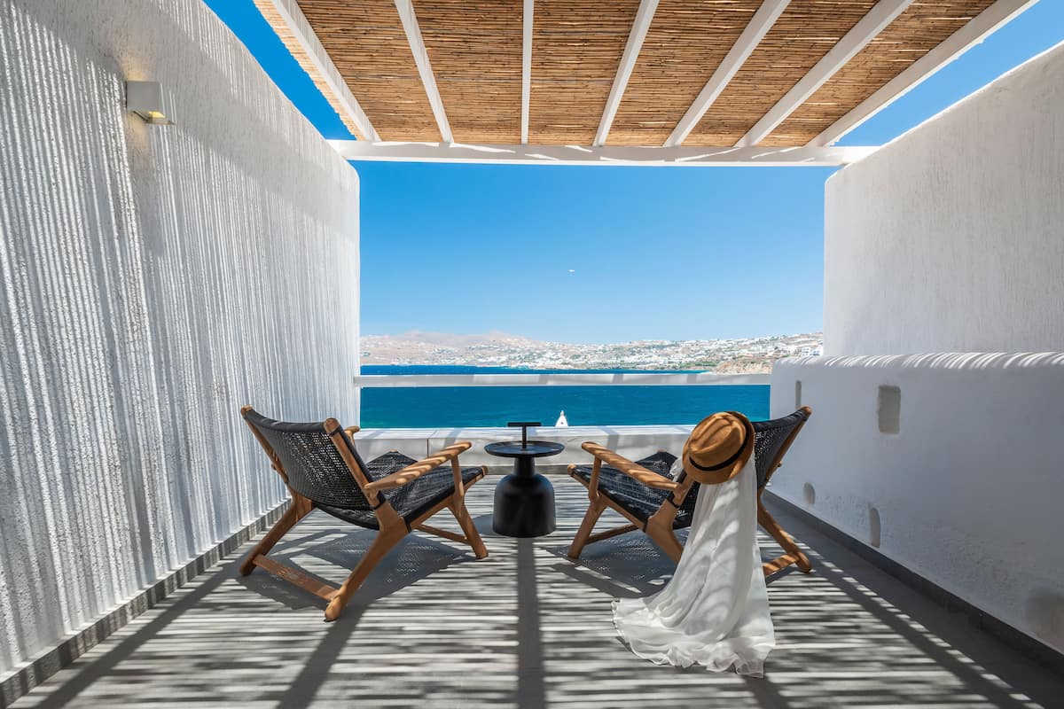 A terrace with a view of the sea, two wicker chairs, a small round table, and a white dress on the floor.