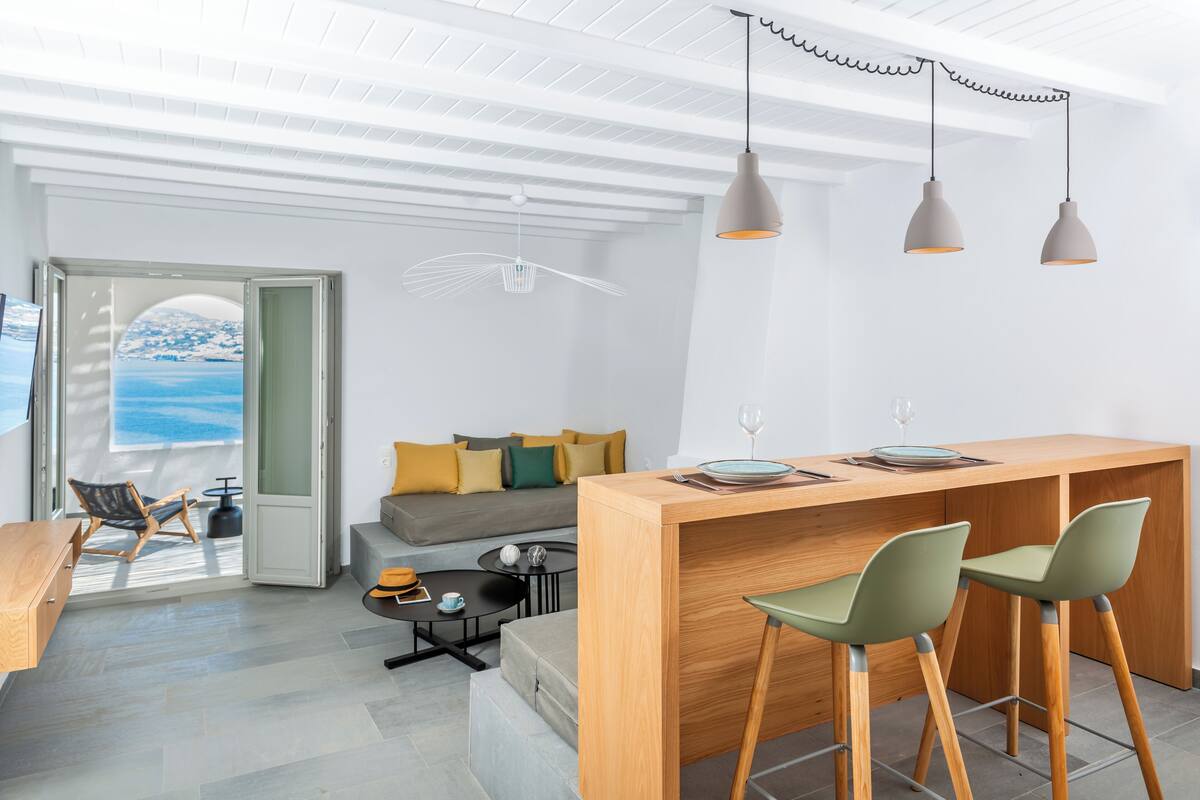 A modern living space with a wooden bar, two bar stools, a sofa with cushions, a dining table, and a view of the sea through an open door.
