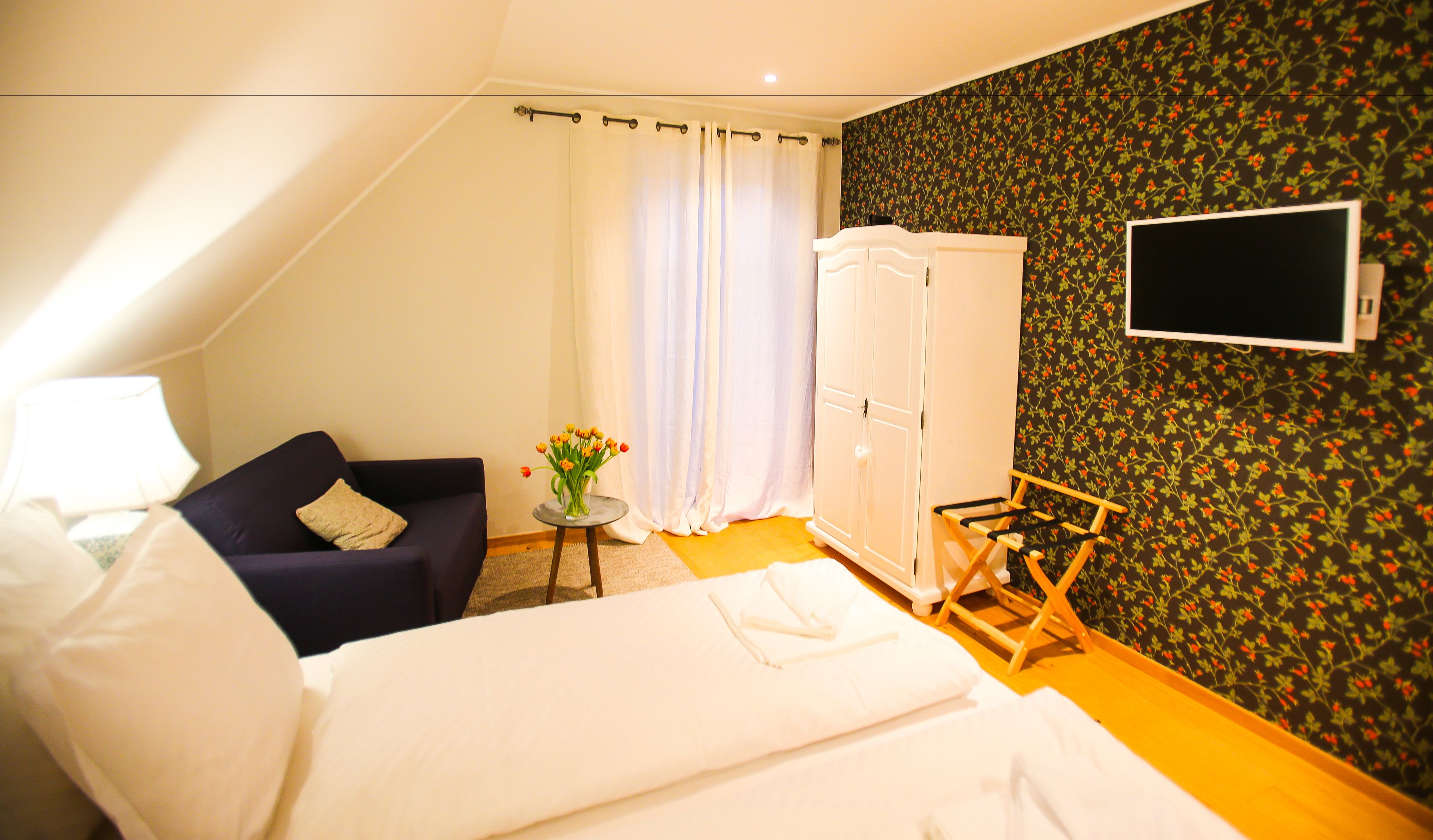 A bedroom with a bed, a sofa, a small table with flowers, a wardrobe, and a TV mounted on the wall.