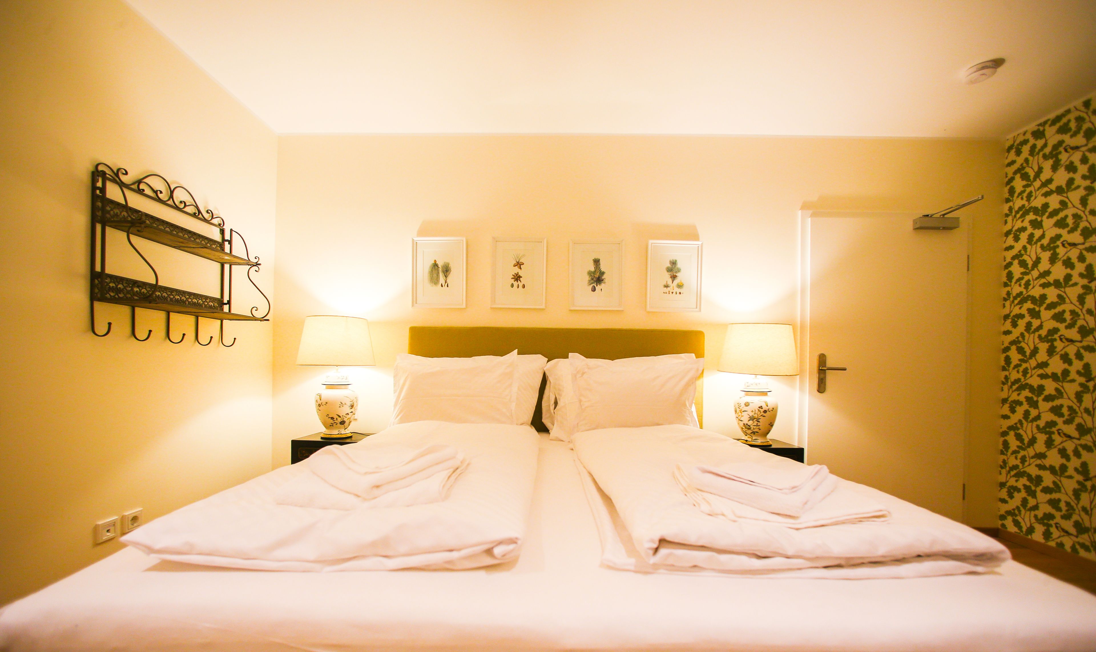 A hotel room with a bed, two bedside tables with lamps, a wall-mounted shelf, and framed botanical prints above the bed.