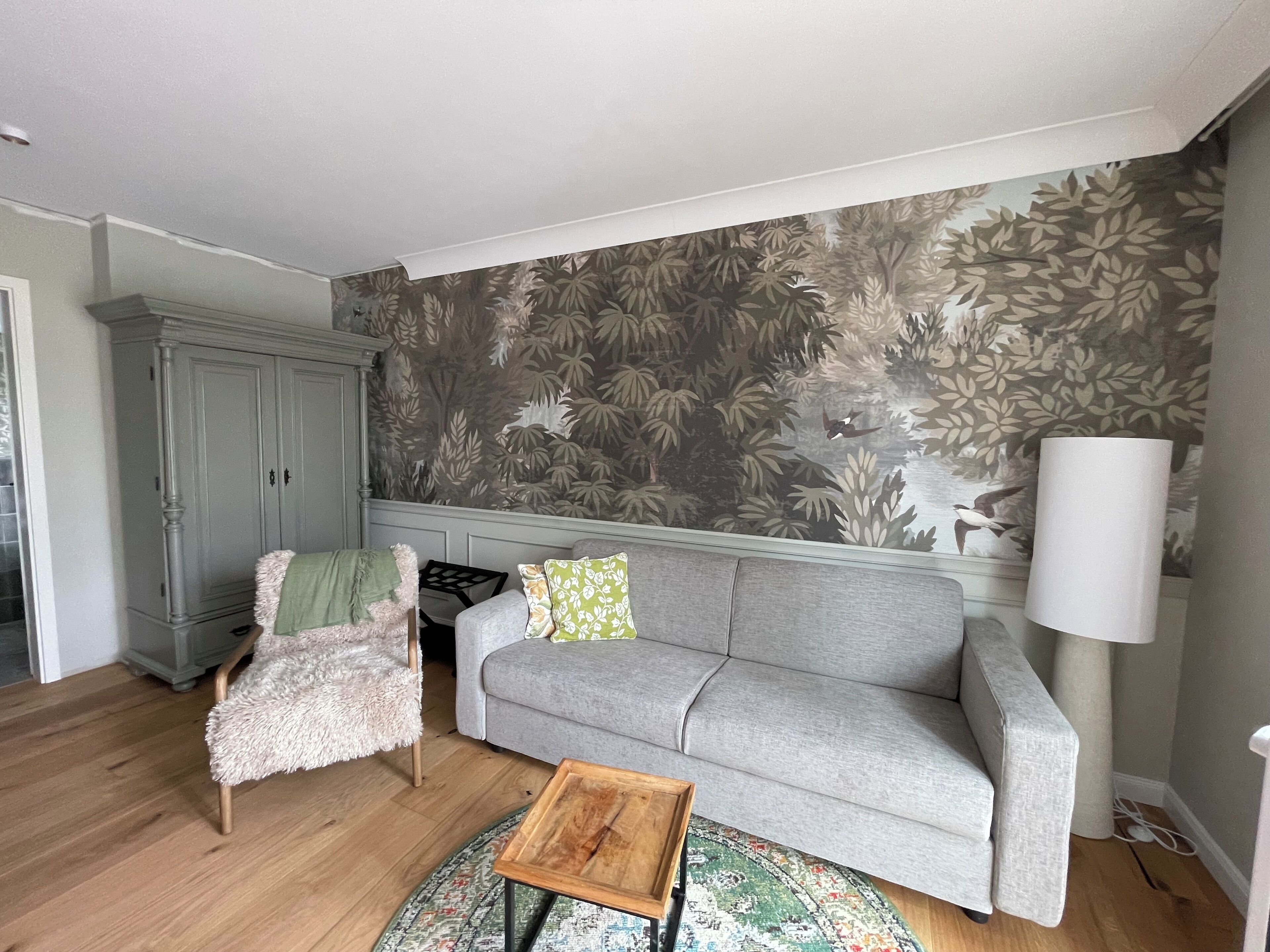 Un salón con un sofá gris, un sillón mullido, una mesa de centro de madera, una alfombra estampada, un mueble de almacenamiento gris y una pared con papel tapiz con motivos vegetales.
