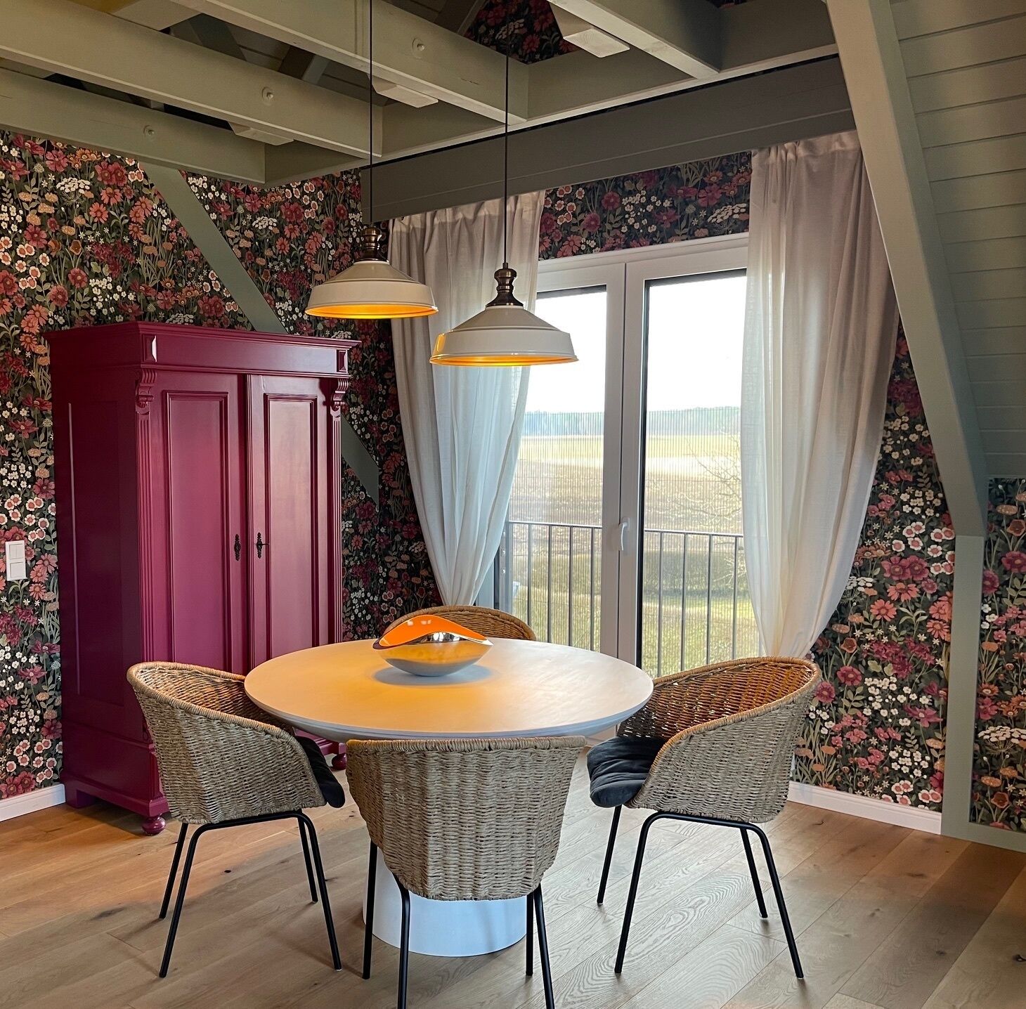 Un comedor con una mesa redonda, sillas de mimbre, un armario rojo y un papel tapiz con motivos florales.