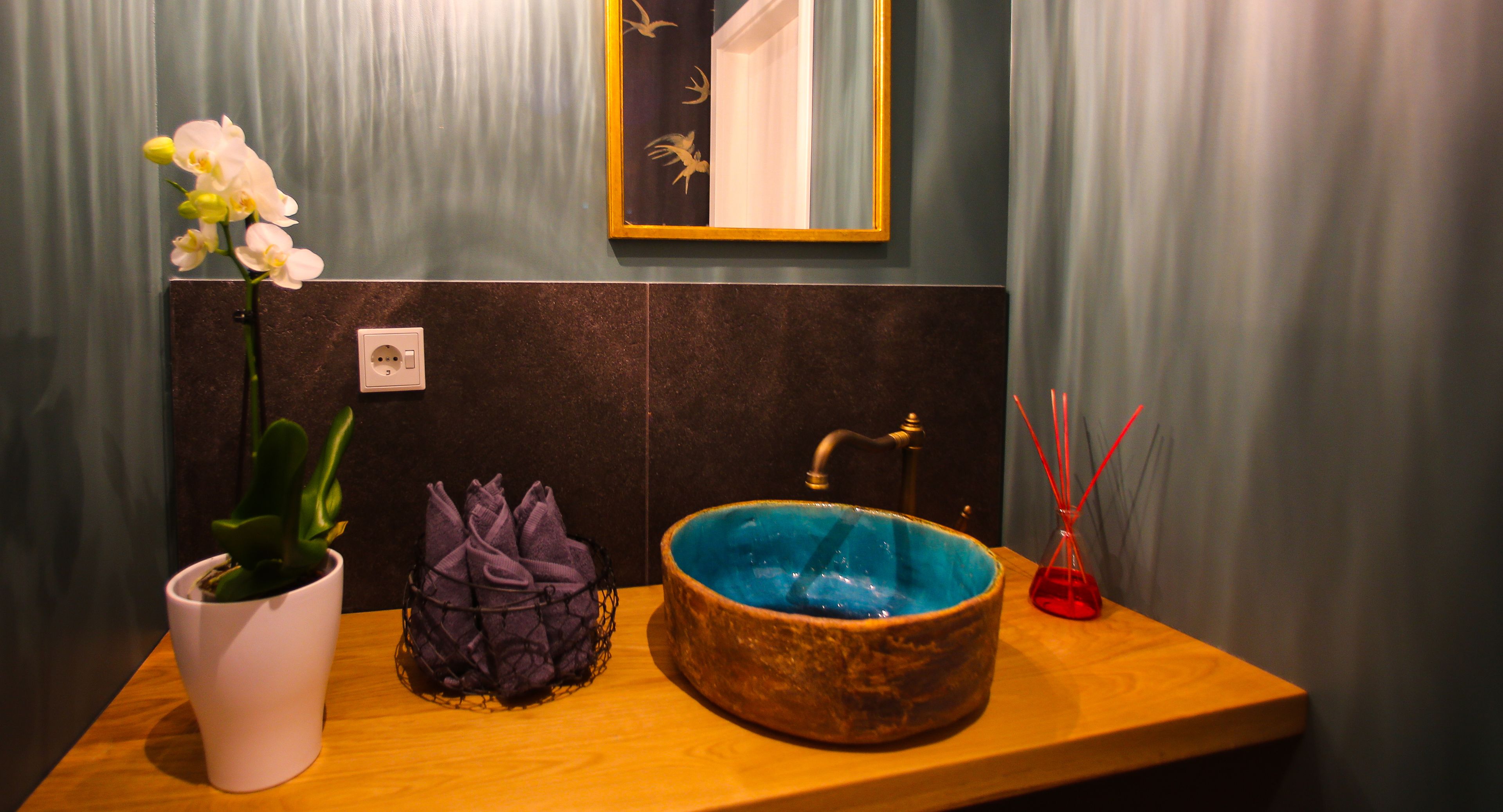 A bathroom with a wooden vanity, a white orchid plant, a purple plant, a blue ceramic sink, and a red glass vase.