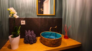 A bathroom with a wooden vanity, a white orchid plant, a purple plant, a blue ceramic sink, and a red glass vase.