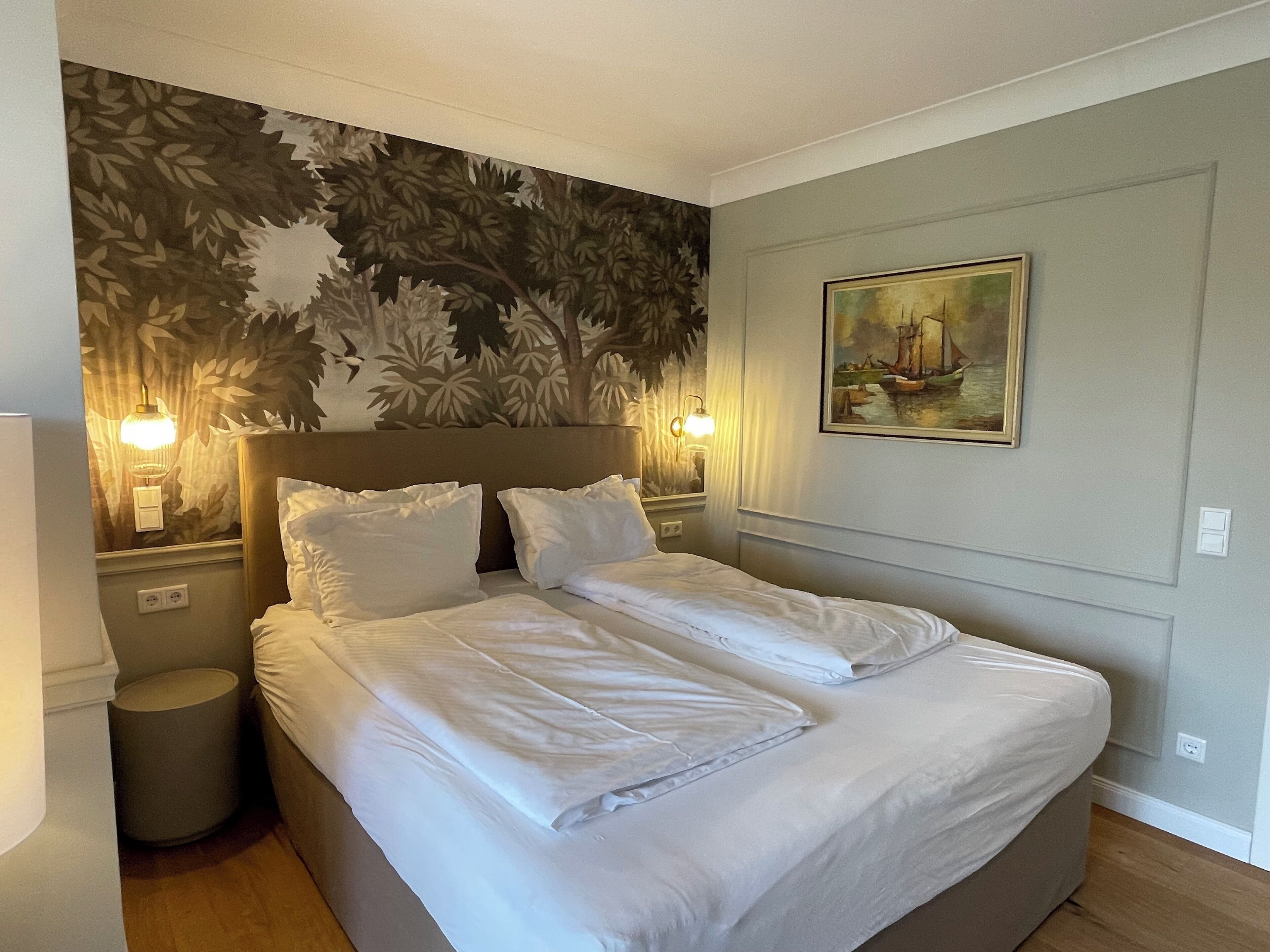 A neatly made bed in a hotel room with a floral wallpaper, two bedside lamps, and a painting on the wall.