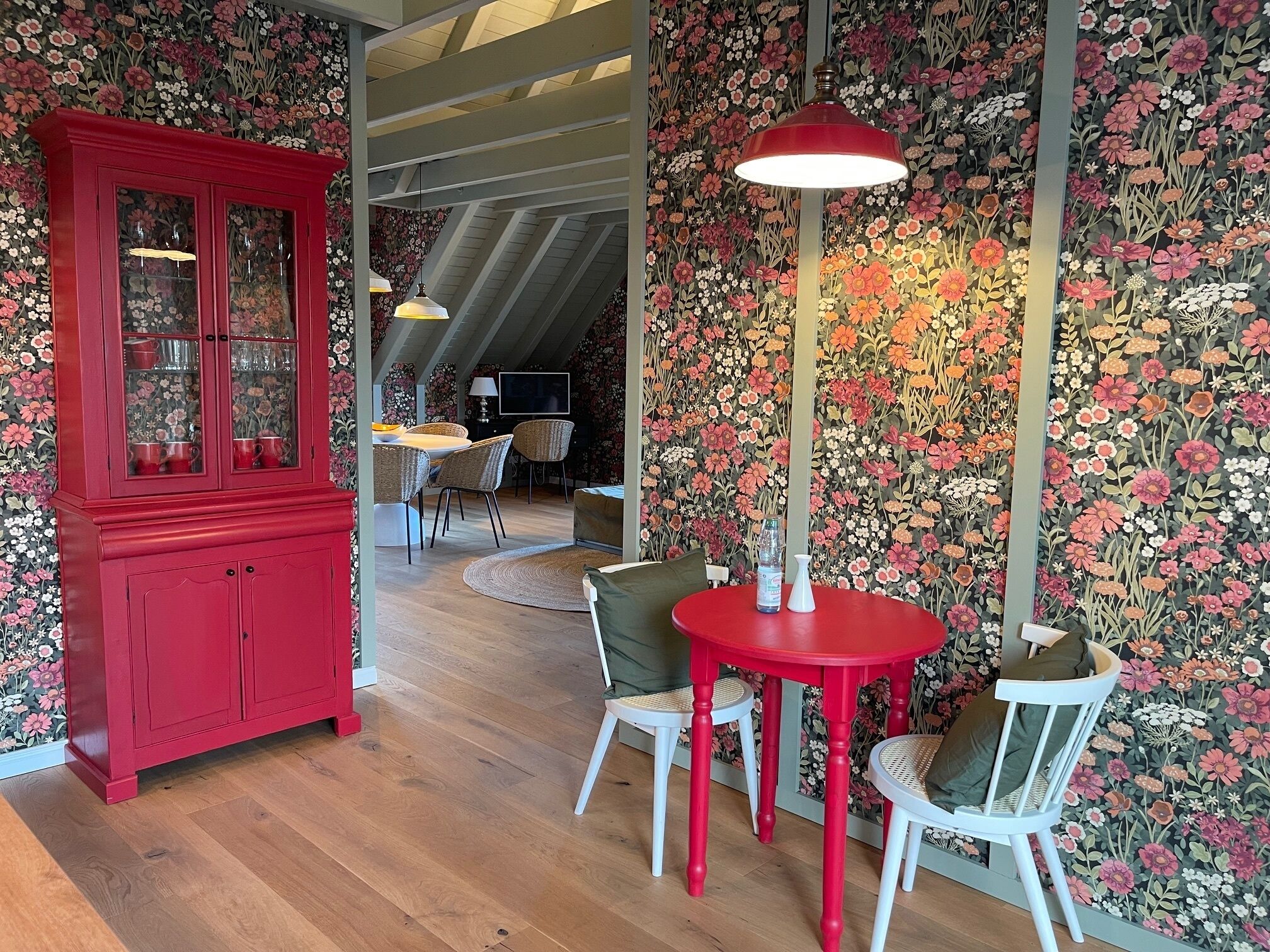A dining area with a red table and chairs, a red cabinet, and floral wallpaper.