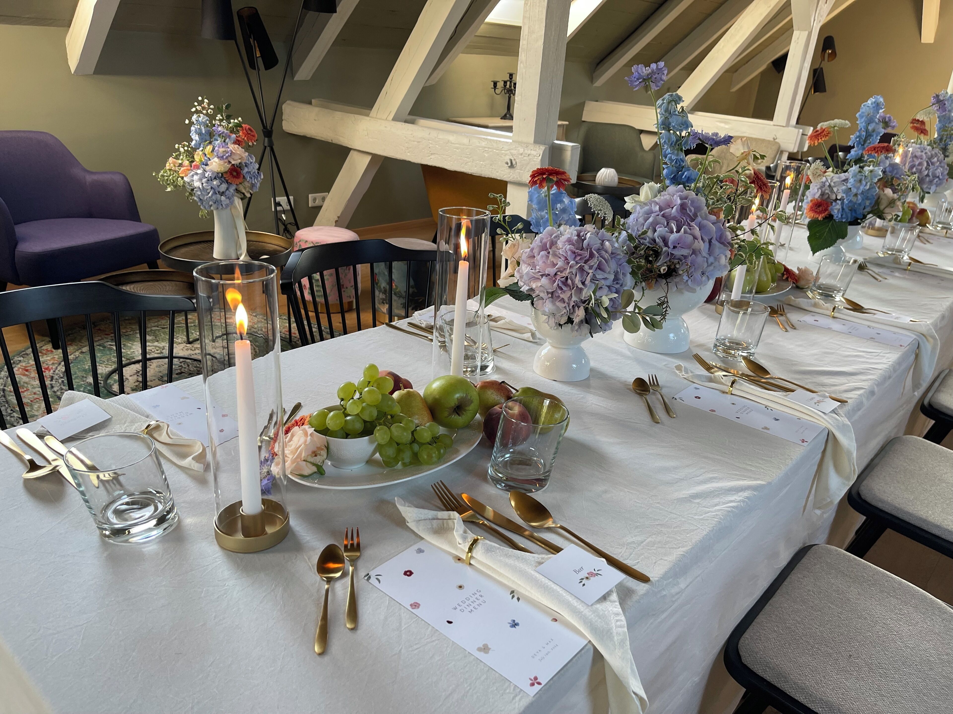 Une longue table à manger, recouverte d’une nappe blanche, agrémentée de couverts dorés et de compositions florales.