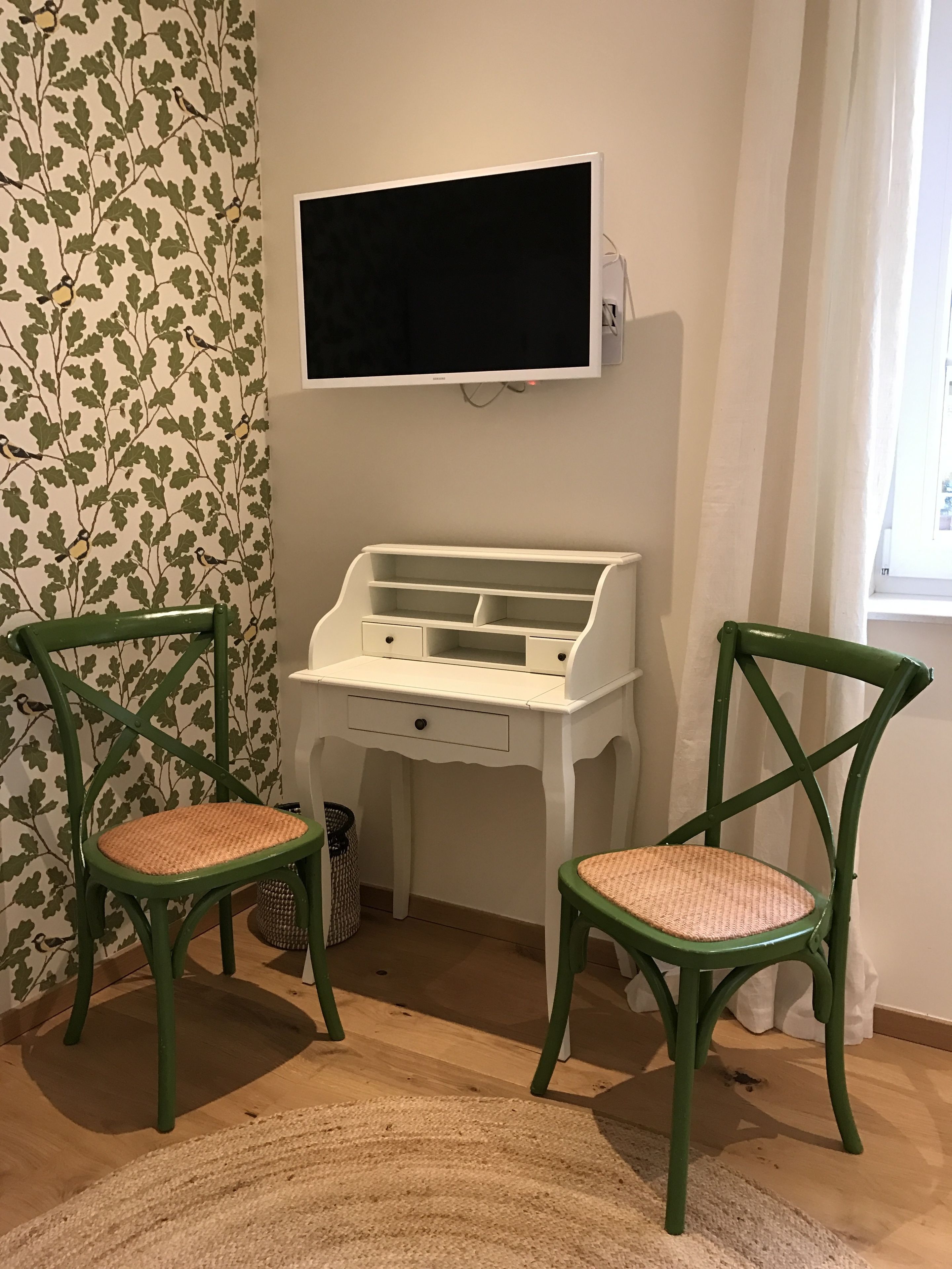 A room with a white desk, two green chairs, a woven rug, and a wall-mounted TV.